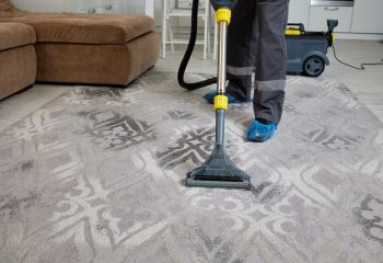 In a wide and bright living room, a diligent worker of a cleaning agency is dressed in overalls, expertly vacuuming and servicing the carpet with precision.